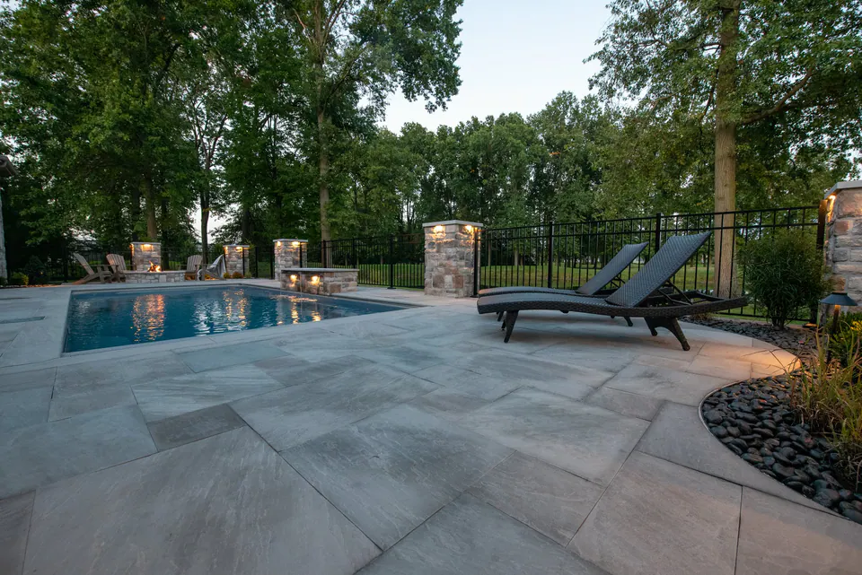 Large bluestone pool patio with lounge chairs, stone piers, black fence, open paving, and planting beds
